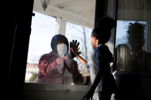 An essential worker in the healthcare industry meets his daughter through a glass window while maintaining a safe distance to protect his family from coronavirus. 